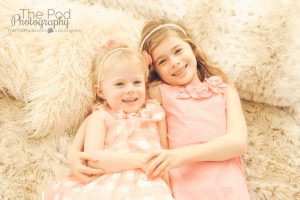two sisters laying on a bed holding hands and looking at the camera, wearing all pink